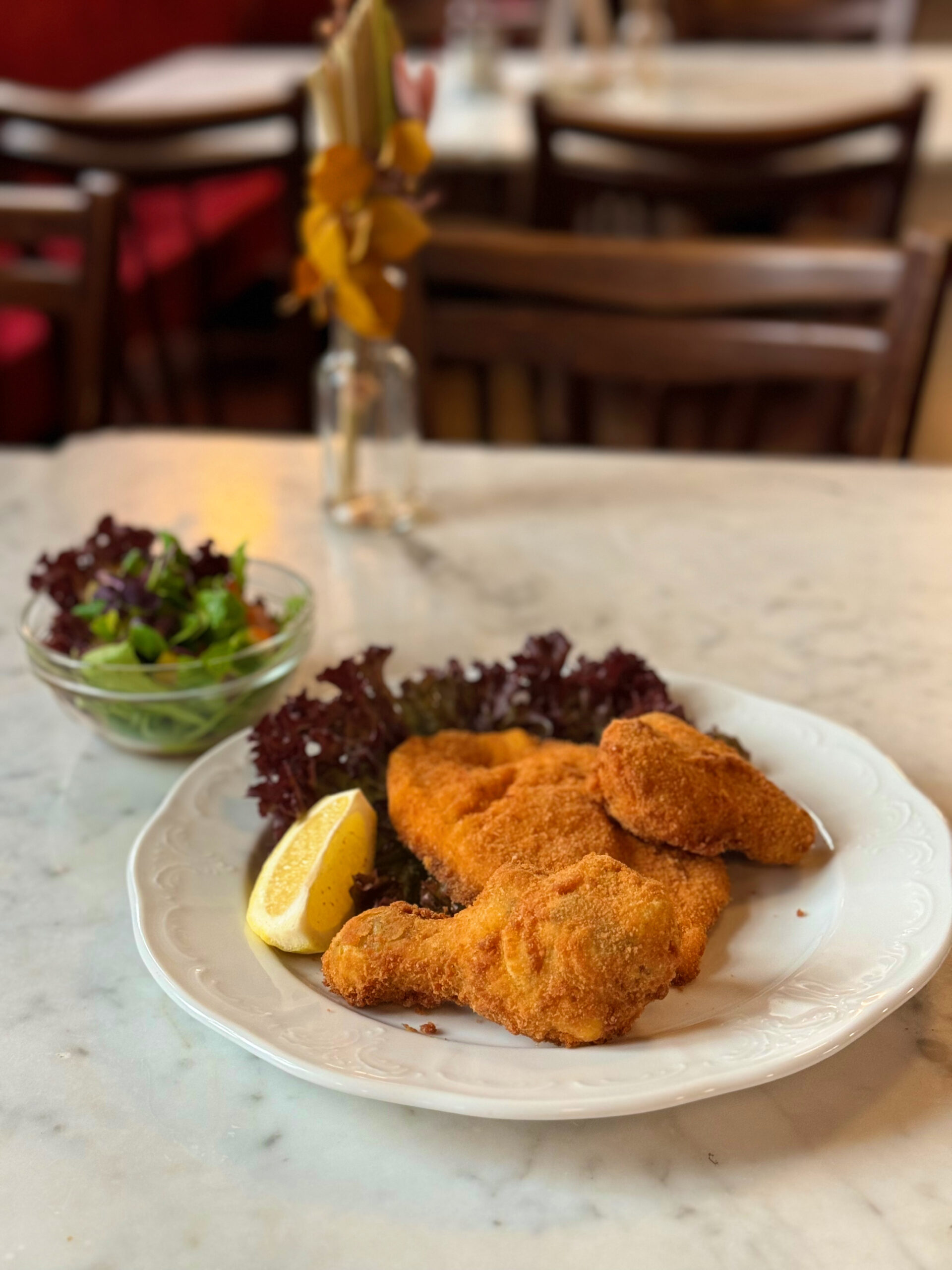 Ein Teller mit Backhendl, serviert mit einer Zitronenscheibe und Salatblättern. Daneben steht eine kleine Schüssel mit gemischtem Salat. Im Hintergrund ist ein gemütlich eingerichtetes Café mit Holzmöbeln und Blumendekorationen zu sehen