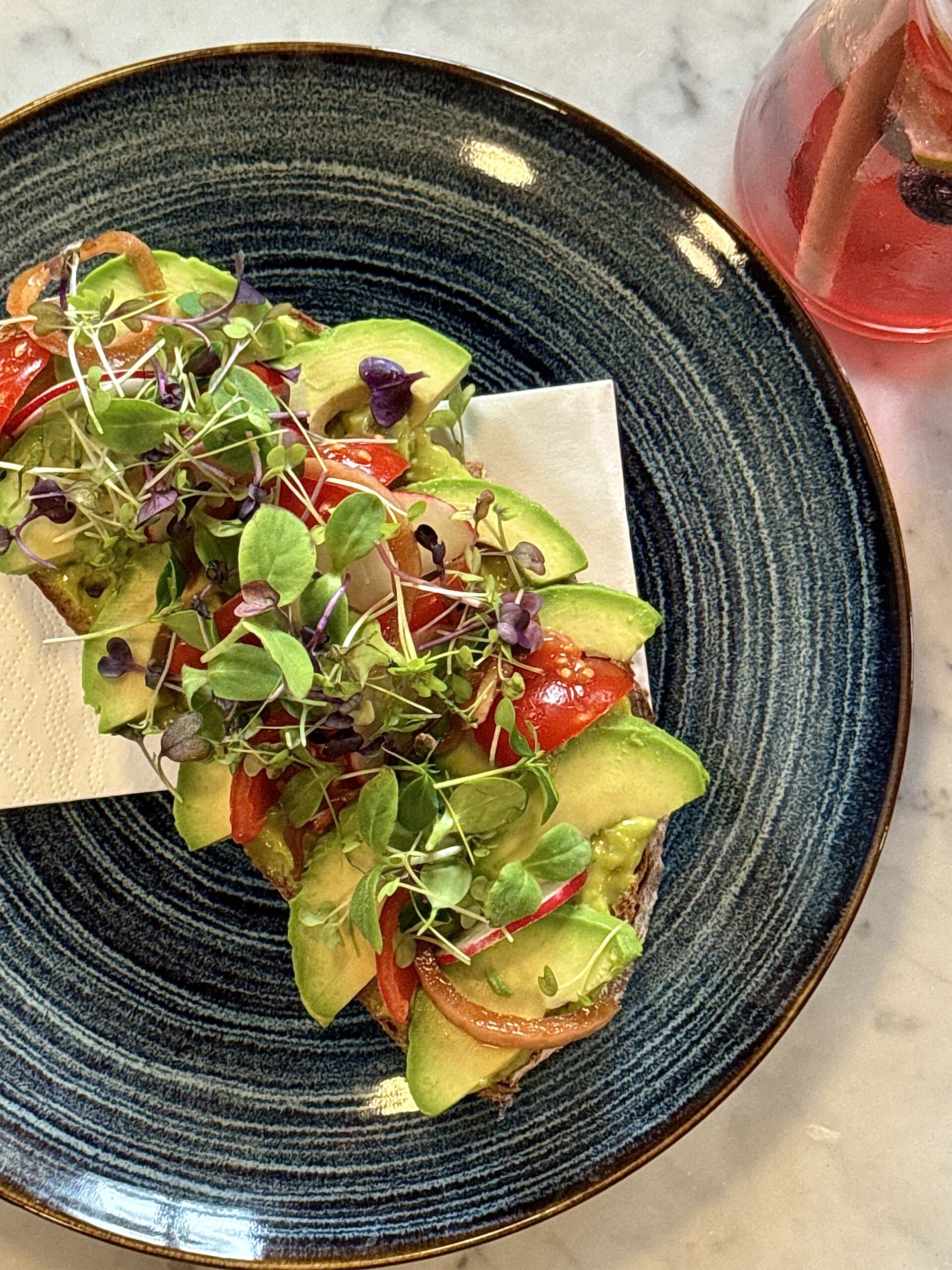 Ein Teller mit Avocado-Toast, garniert mit frischen Tomaten, roten Zwiebeln und einer großzügigen Portion Microgreens. Im Hintergrund ist ein Glas mit einem erfrischenden Getränk zu sehen.