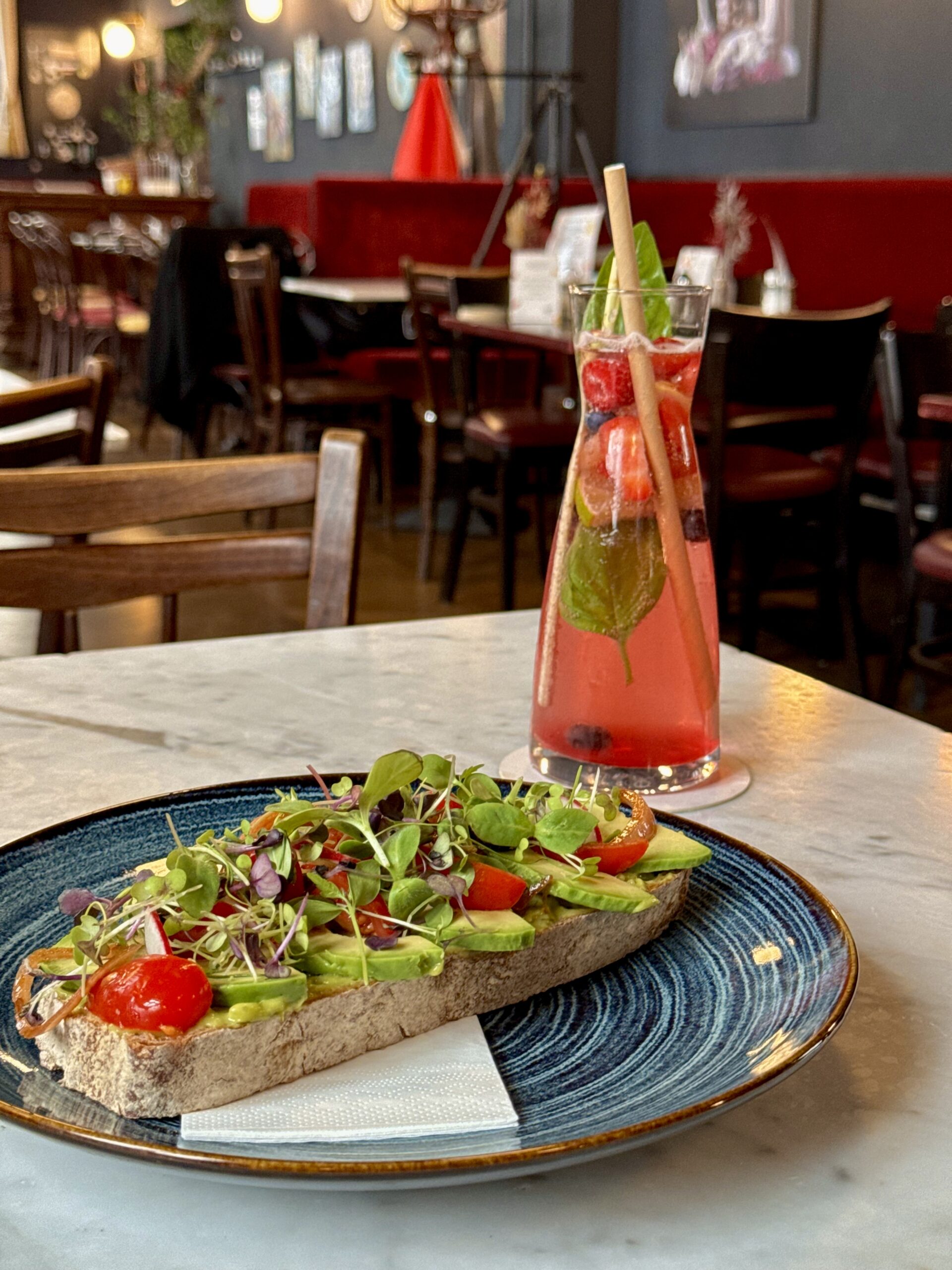 Ein Teller mit Avocado-Toast, garniert mit frischen Tomaten und Microgreens, serviert auf einem Tisch in einem gemütlichen Café. Daneben steht ein großes Glas mit einem erfrischenden roten Getränk, dekoriert mit frischen Minzblättern. Im Hintergrund ist das stilvoll eingerichtete Café mit roten Sitzbänken und hölzernen Stühlen zu sehen.