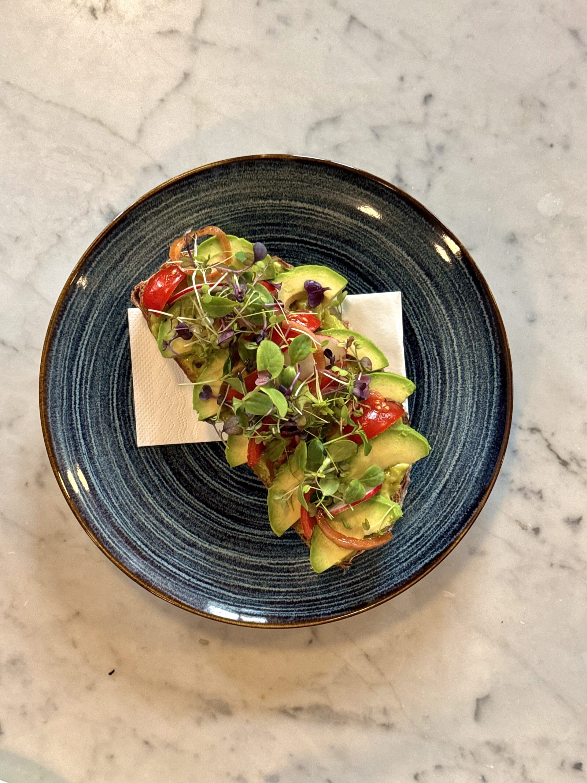 Ein Teller mit Avocado-Toast, garniert mit frischen Tomaten, roten Zwiebeln und einer großzügigen Portion Microgreens. Im Hintergrund ist ein Glas mit einem erfrischenden Getränk zu sehen.