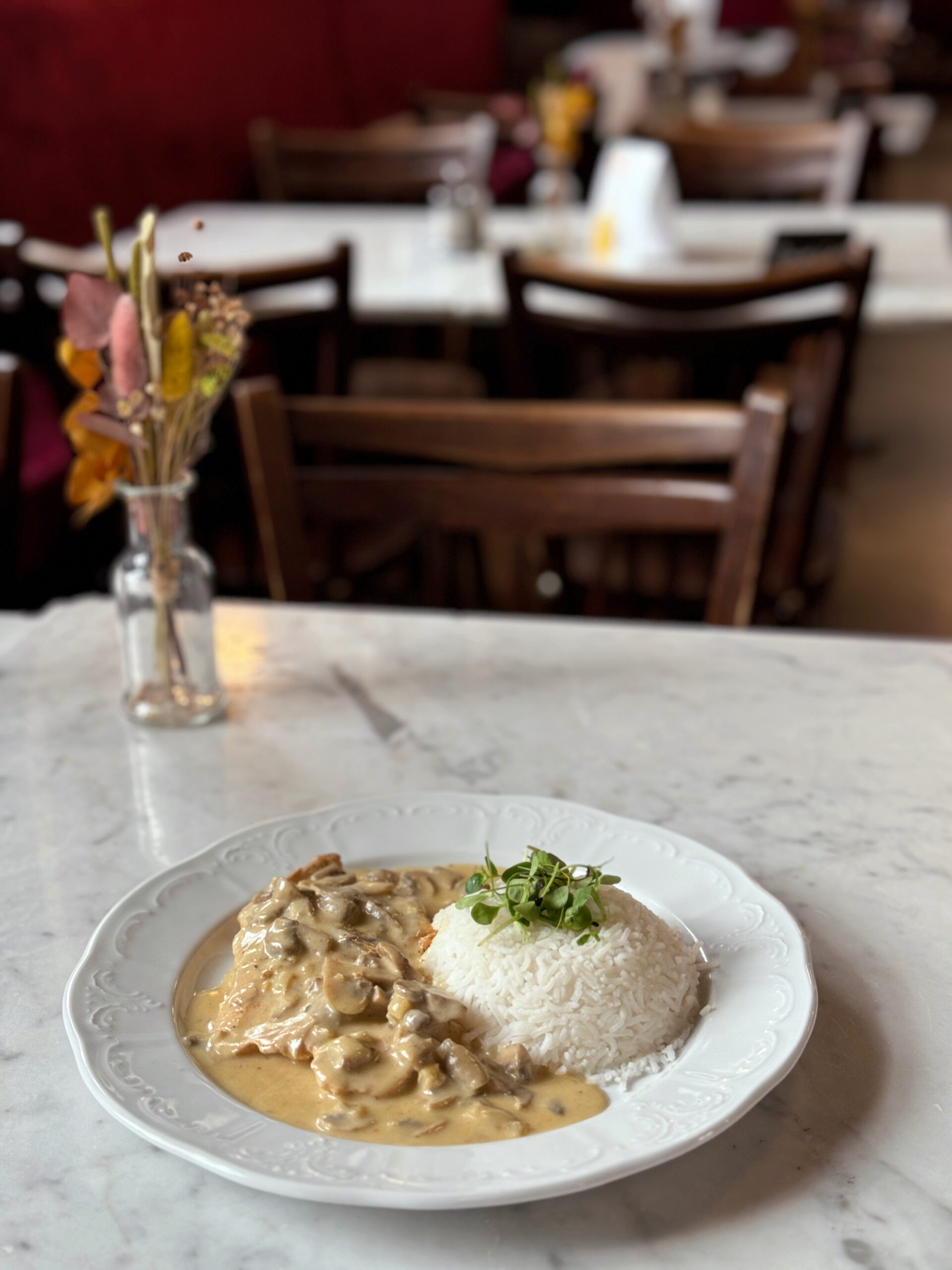 Ein Teller mit Putenbrustfilet in einer cremigen Champignonsauce, serviert mit Reis und garniert mit frischen Kräutern. Im Hintergrund ist ein gemütlich eingerichtetes Café mit Holzmöbeln und Blumendekorationen zu sehen.